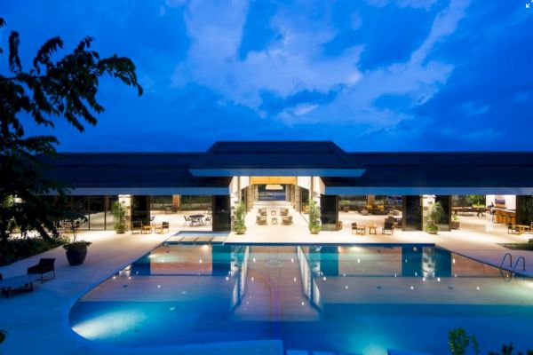 A luxurious, modern building with large windows is shown at dusk, featuring a well-lit swimming pool surrounded by lounge chairs.