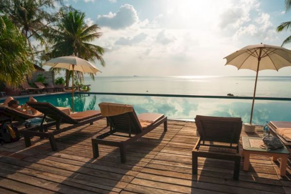 A serene poolside with lounge chairs, umbrellas, palm trees, and a beautiful ocean view under a partly cloudy sky.