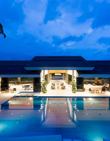 A luxury building with an illuminated swimming pool, surrounded by trees, under a dramatic blue evening sky.