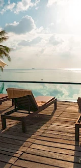 A serene poolside scene with lounge chairs and umbrellas overlooks a calm sea under a partly cloudy sky at sunset or sunrise.