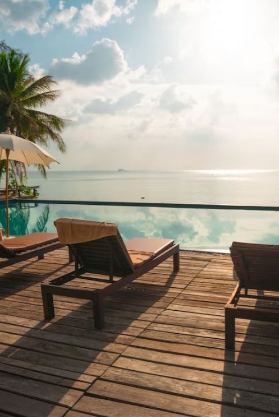A serene poolside setting with lounge chairs, umbrellas, and palm trees, overlooking a calm body of water under a partly cloudy sky.