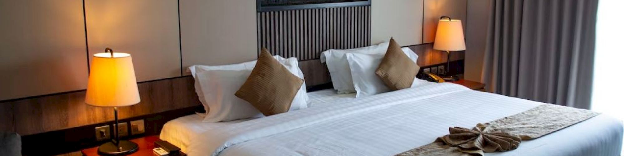 A cozy hotel room with a large bed, decorative pillows, lamps on nightstands, and draped curtains by the window, creating a warm ambiance.