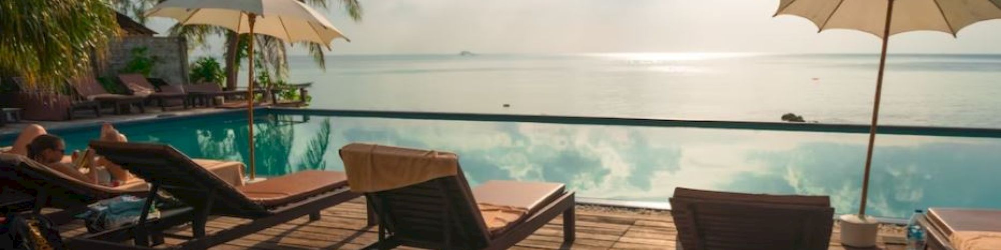 A serene poolside scene with lounge chairs, umbrellas, and palm trees, overlooking calm water under a partly cloudy sky.