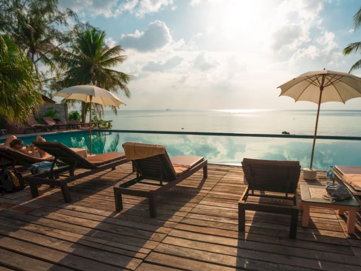 Lounge chairs and umbrellas are set by a pool, overlooking a serene ocean view with palm trees and a partly cloudy sky.
