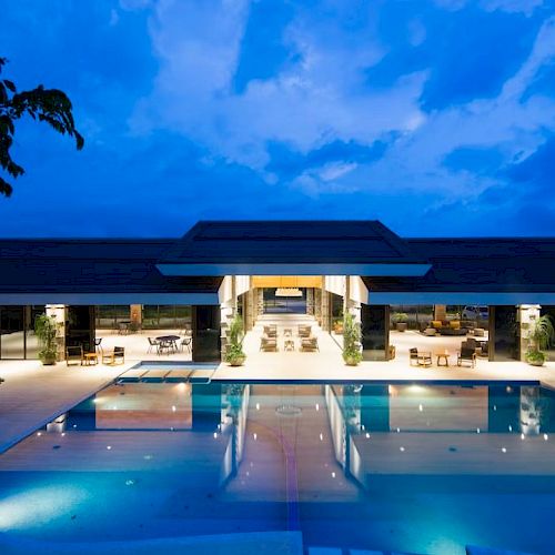 A luxurious modern building with a large illuminated pool in the foreground, surrounded by seating areas under a twilight sky.