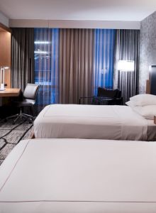 The image shows a modern hotel room with two beds, a TV, a desk, and contemporary decor.