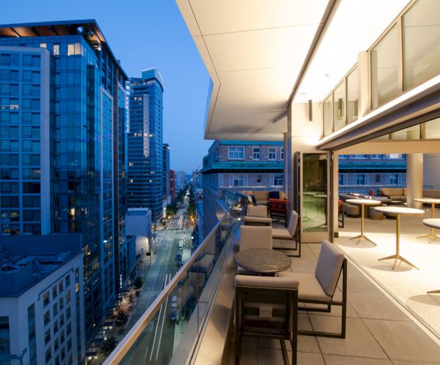 Rooftop terrace with seating overlooks a city street lined with modern skyscrapers at dusk.