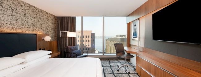 A modern hotel room with a large bed, flat-screen TV, desk, and window view of city buildings and water.