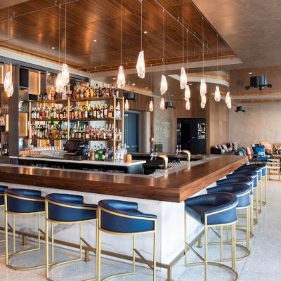 The image shows a modern bar with overhanging lights, a stocked liquor shelf, and seating area with blue chairs and lounge seats.