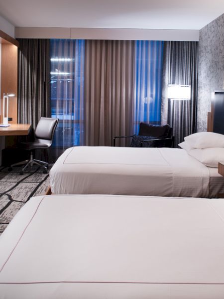 A modern hotel room with two beds, a flat-screen TV, desk, chair, and stylish lighting and decor.