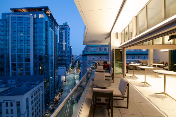 The image shows a modern balcony with seating, overlooking a cityscape at dusk, with tall buildings and streetlights illuminating the scene.