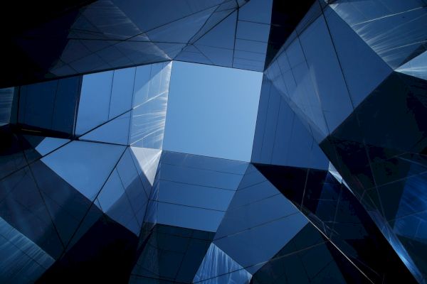 The image shows an upward view of geometric, mirrored buildings surrounding a clear blue sky, creating a modern architectural pattern.