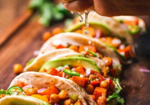 A row of vegetable tacos on a wooden board with a hand squeezing lime juice over them, garnished with cilantro and lime slices.