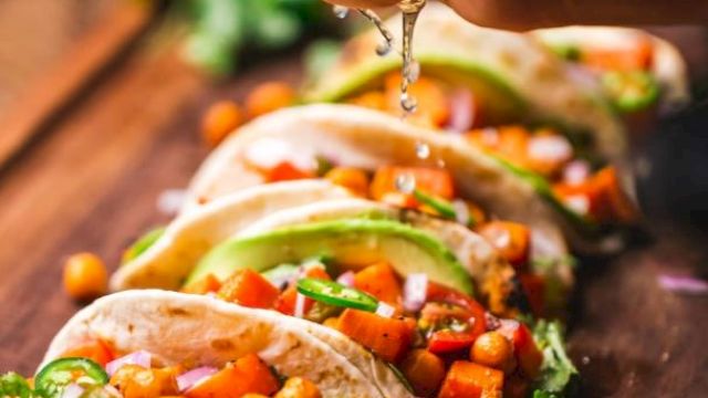 A row of vegetable tacos on a wooden board with a hand squeezing lime juice over them, garnished with cilantro and lime slices.