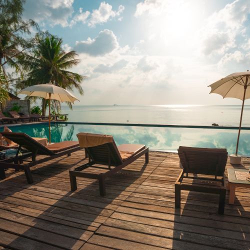Sunlit poolside deck with lounge chairs, umbrellas, palm trees, and ocean view under a partly cloudy sky.