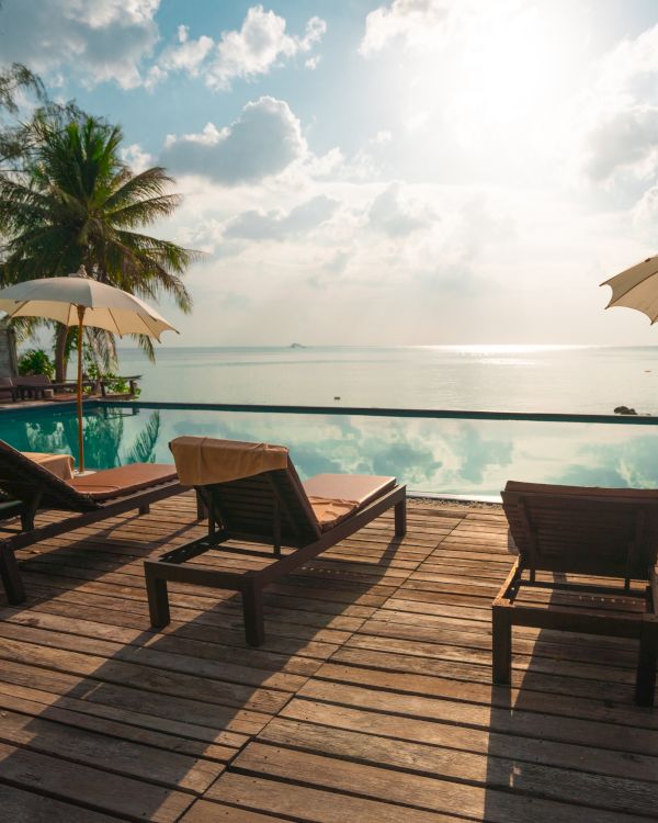 Sunlit poolside deck with lounge chairs, umbrellas, palm trees, and ocean view under a partly cloudy sky.