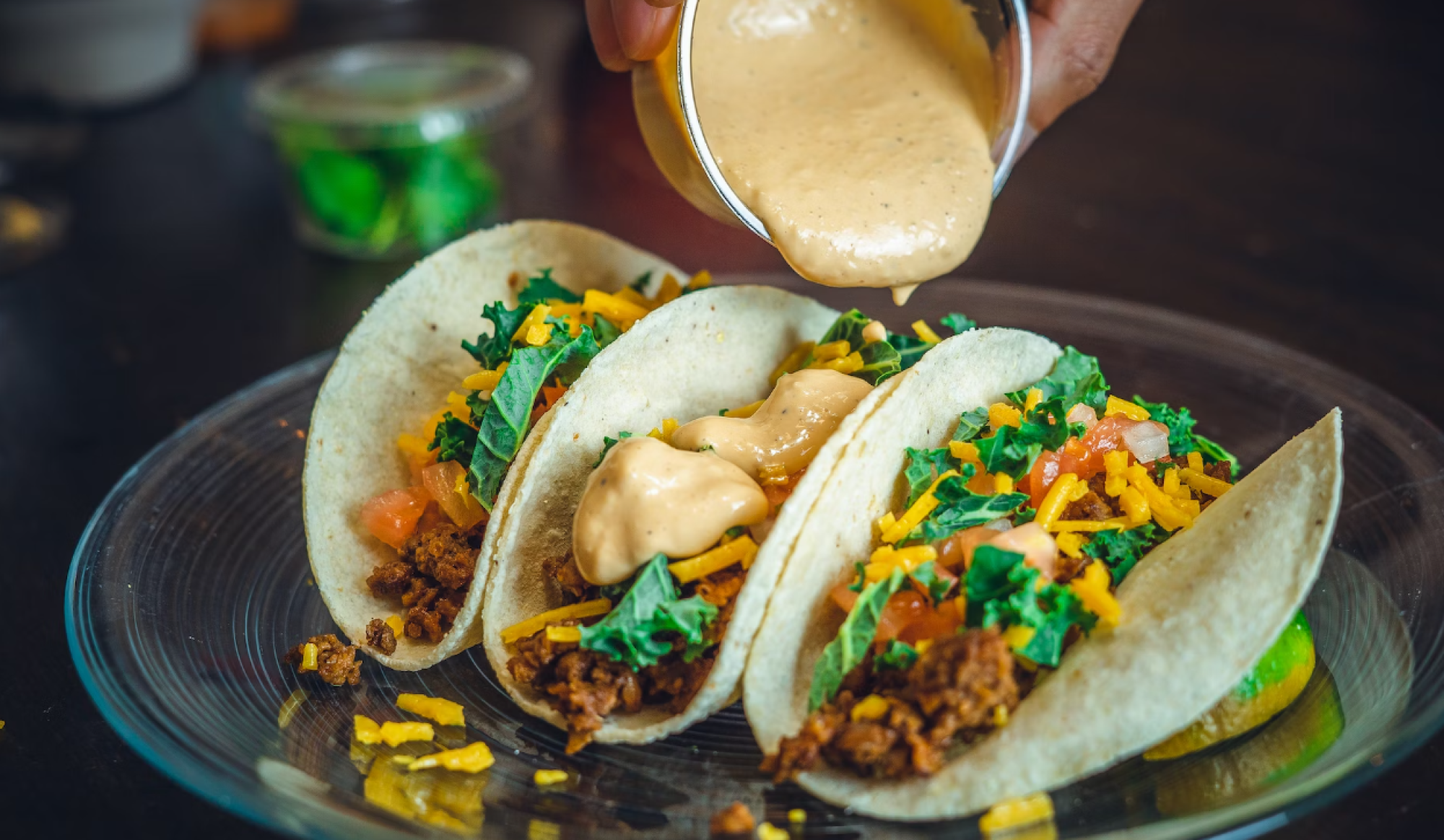 Three tacos filled with toppings like meat, cheese, and greens, being drizzled with creamy sauce on a plate.