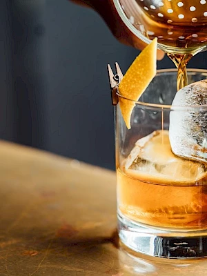A cocktail being poured over ice in a glass with a citrus twist garnish, on a bar counter.