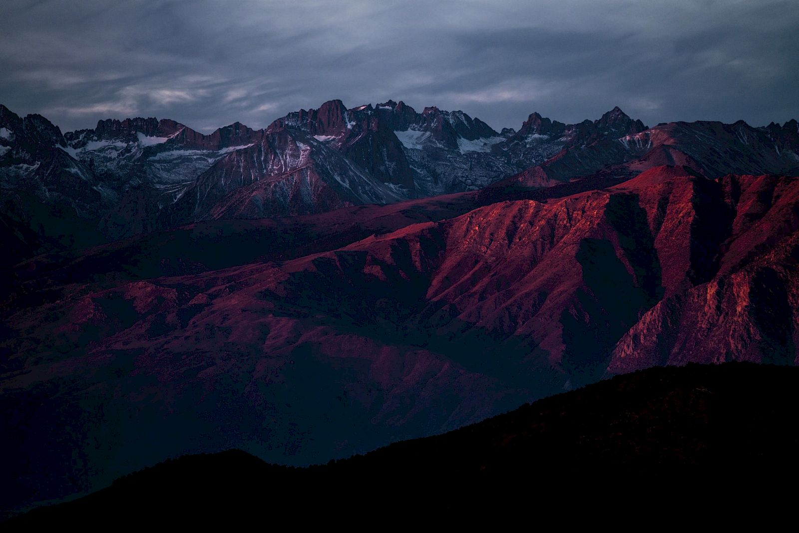 A mountain range at sunset, with peaks and ridges bathed in a reddish hue under a dramatic, cloudy sky.