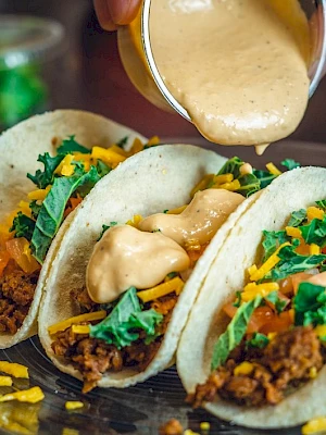 Three tacos filled with meat, cheese, lettuce, and tomatoes, with sauce being poured on top, presented on a glass plate.