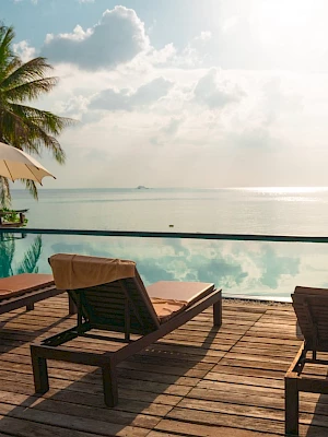 Poolside view with lounge chairs, umbrellas, and a serene ocean background, surrounded by palm trees under a partly cloudy sky.