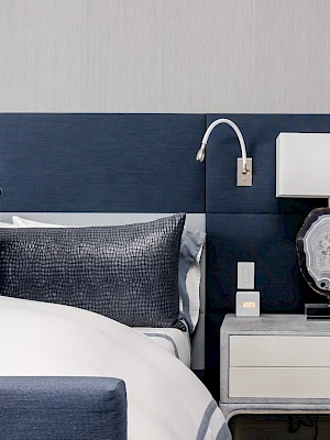 A stylish bedroom with a blue headboard, modern lamp, and cozy bedding. A nightstand with drawers and decorative elements completes the look.