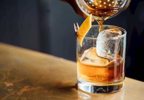 A cocktail is being poured into a glass with ice and a lemon twist garnish on a bar counter.