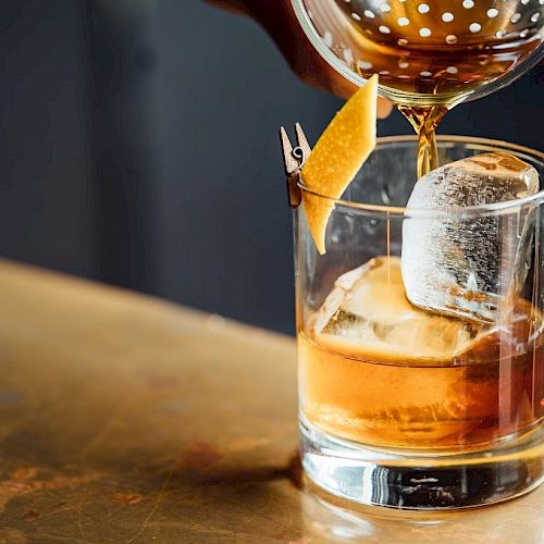 A cocktail is being poured into a glass with ice and a lemon twist garnish on a bar counter.