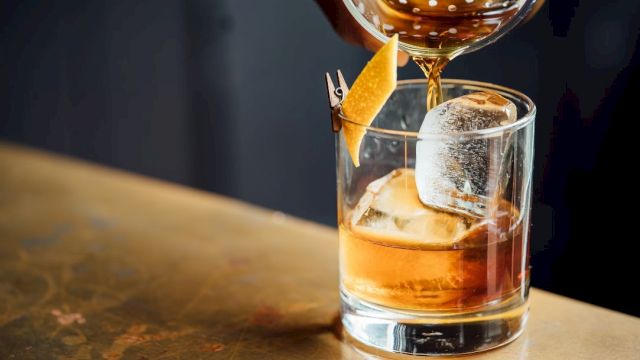 A cocktail is being poured into a glass with ice and a lemon twist garnish on a bar counter.