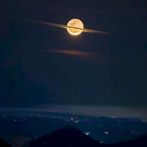 A full moon appears to have rings like Saturn due to cloud formations, set against a night sky with distant city lights.