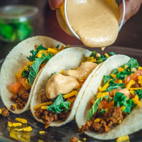 Three tacos with meat, cheese, veggies, and sauce being poured, served on a glass plate.