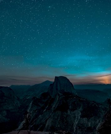 A mountainous landscape is silhouetted against a starry night sky with a glowing horizon and wispy clouds above.
