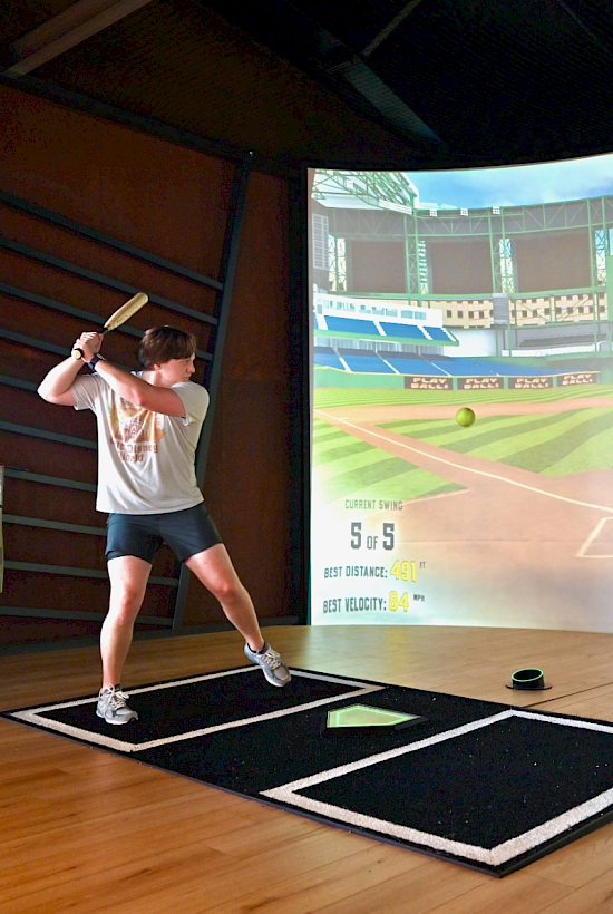 A person is playing a virtual baseball game, swinging a bat at a digital pitch on a large screen indoors.