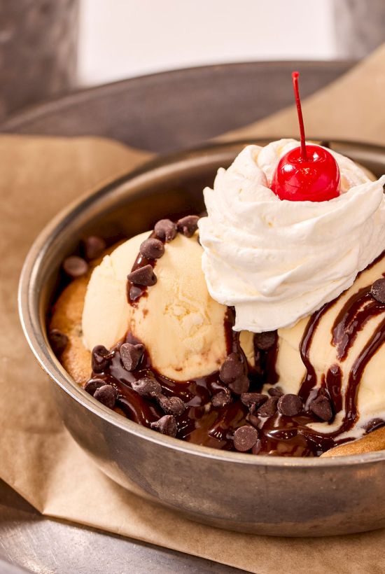 A skillet cookie topped with vanilla ice cream, chocolate syrup, whipped cream, and a cherry, served on a piece of parchment paper.