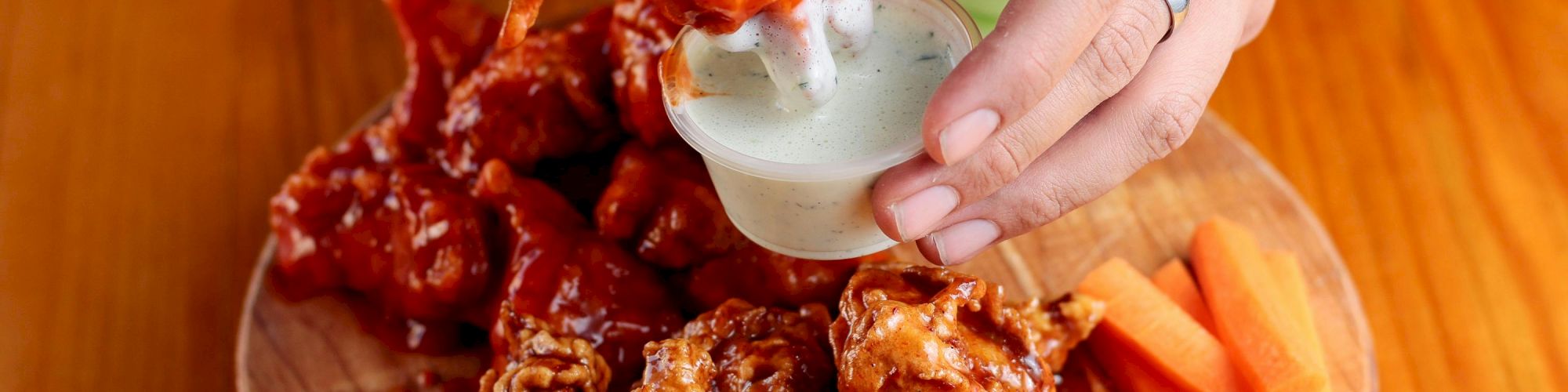 A person dipping a saucy chicken wing into a white sauce, with more wings and carrot sticks on a wooden platter.