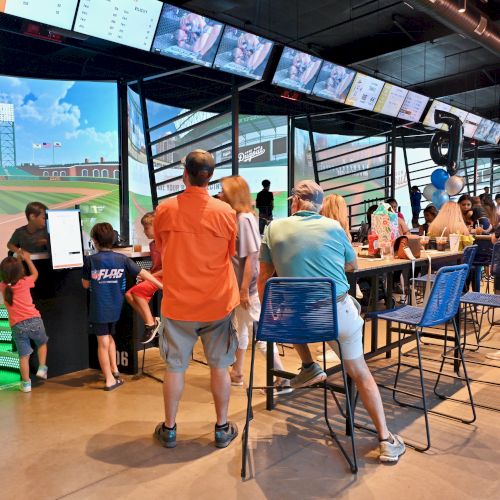 People gathered indoors, watching a sports simulation on a large screen. Some are sitting, others standing, in a lively atmosphere.