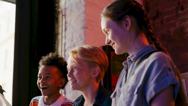 Three smiling kids are inside, looking towards something with expressions of wonder or joy. They are captured in natural light and casual attire.