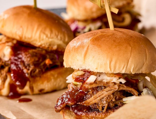 Three pulled pork sliders with barbecue sauce and coleslaw on a bun, held together with skewers, placed on a slate plate.