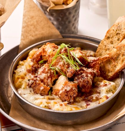 A bowl of creamy mac and cheese topped with crispy chicken, garnished with herbs, served with bread slices and popcorn in the background.