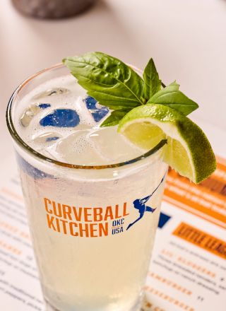 A refreshing beverage with ice, basil, and a lime slice in a glass labeled "Curveball Kitchen OKC USA" on a menu background.