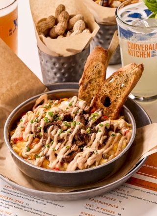 A dish with toppings and toast, peanuts in a container, and a drink with a lemon slice on a table with menus underneath.