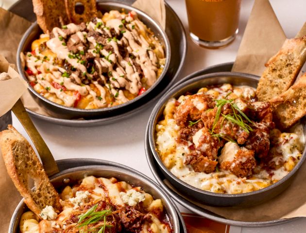 The image shows three bowls of mac 'n cheese topped with various ingredients, bread slices, a glass of beer, and a container of popcorn.