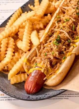 The image shows a hot dog topped with crispy onions and herbs, served with crinkle-cut fries on a plate.