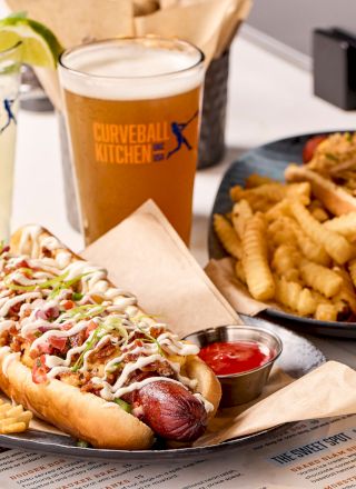 Plates with hot dogs topped with sauces, crinkle-cut fries, a pint of beer, and a lemonade are on a table.