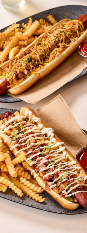 Two loaded hot dogs with crinkle-cut fries on plates, served with ketchup and a pint of beer.