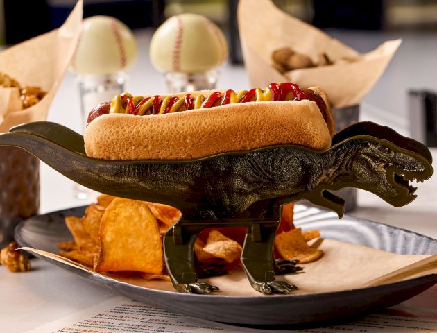 A toy dinosaur creatively holds a hot dog on its back, surrounded by snacks like chips, in a playful food presentation.