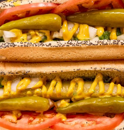 Two hot dogs in poppy seed buns topped with mustard, pickles, onions, tomatoes, and relish. Crinkle-cut fries are in the background.
