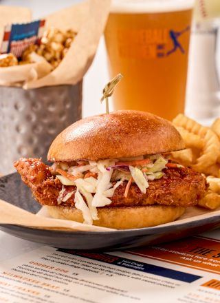 A chicken sandwich with coleslaw, fries, popcorn, and a beer are served on a table with a menu in the background.