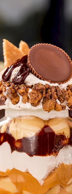 A dessert in a jar layered with whipped cream, chocolate sauce, and topped with a peanut butter cup and cookies.