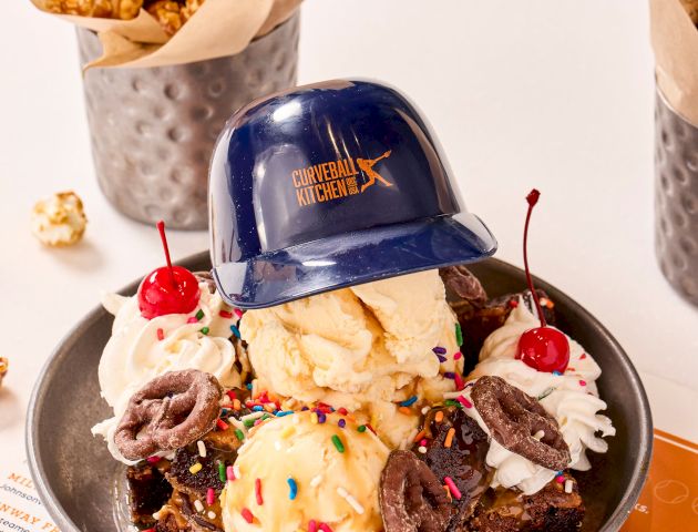 A dessert with ice cream, cherries, and sprinkles in a bowl. Topped with a small helmet, next to popcorn in metal cups.